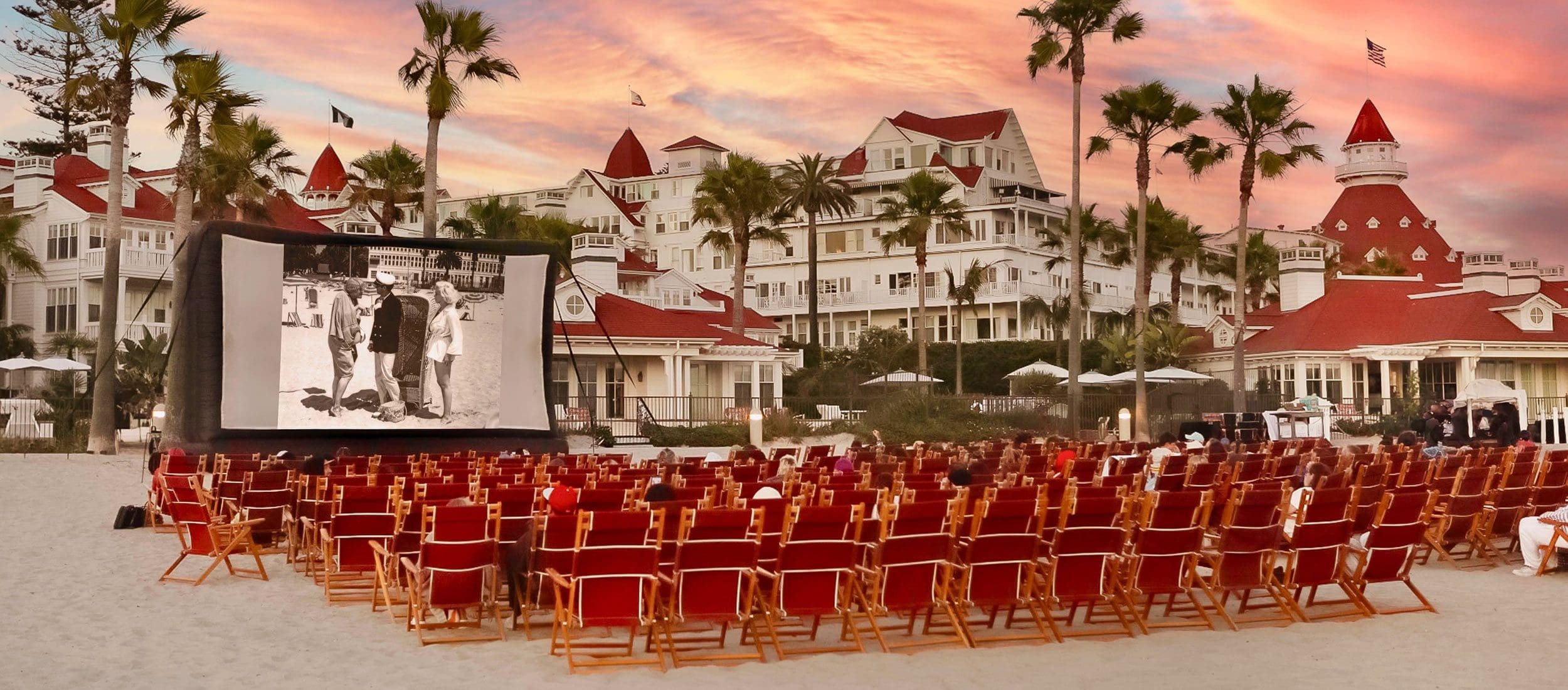Rows of red beach chairs face an outdoor movie screen in front of Hotel del Coronado at sunset.